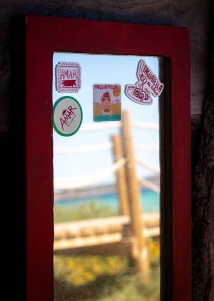 Un marco de ventana pintado de rojo y cubierto de pegatinas de AMAR, con vistas al mar turquesa.