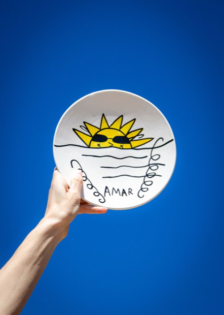 Una mano sujetando en alto un plato color crema pintado con un sol con gafas, sobre un fondo azul intenso.