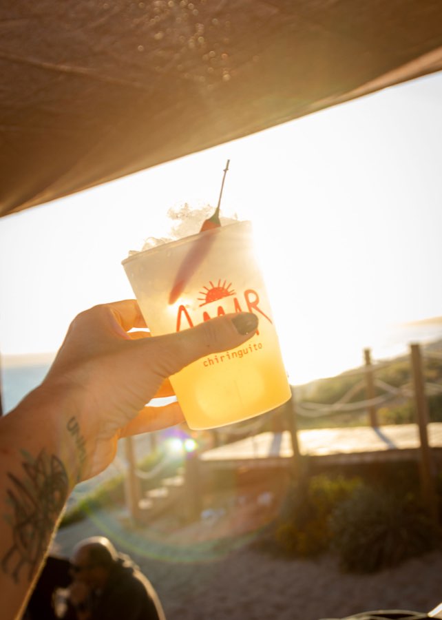 Una mano levantando un vaso de AMAR contra el sol poniente, con la terraza en silueta detrás.