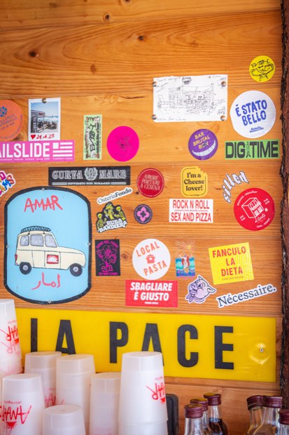 Una pared de madera cubierta de pegatinas sobre un estante con vasos de AMAR apilados.