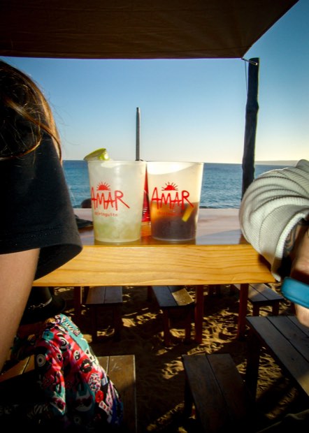Dos vasos de AMAR con bebidas con hielo sobre una mesa de madera, con el mar y el sol bajo detrás.
