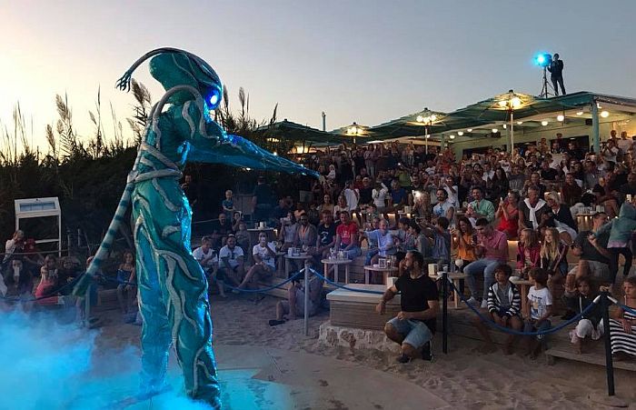 El Alienshow en Blue Bar: un artista con un traje de criatura turquesa que brilla, de pie entre humo azul sobre la arena, ante el p&uacute;blico en las gradas al anochecer.