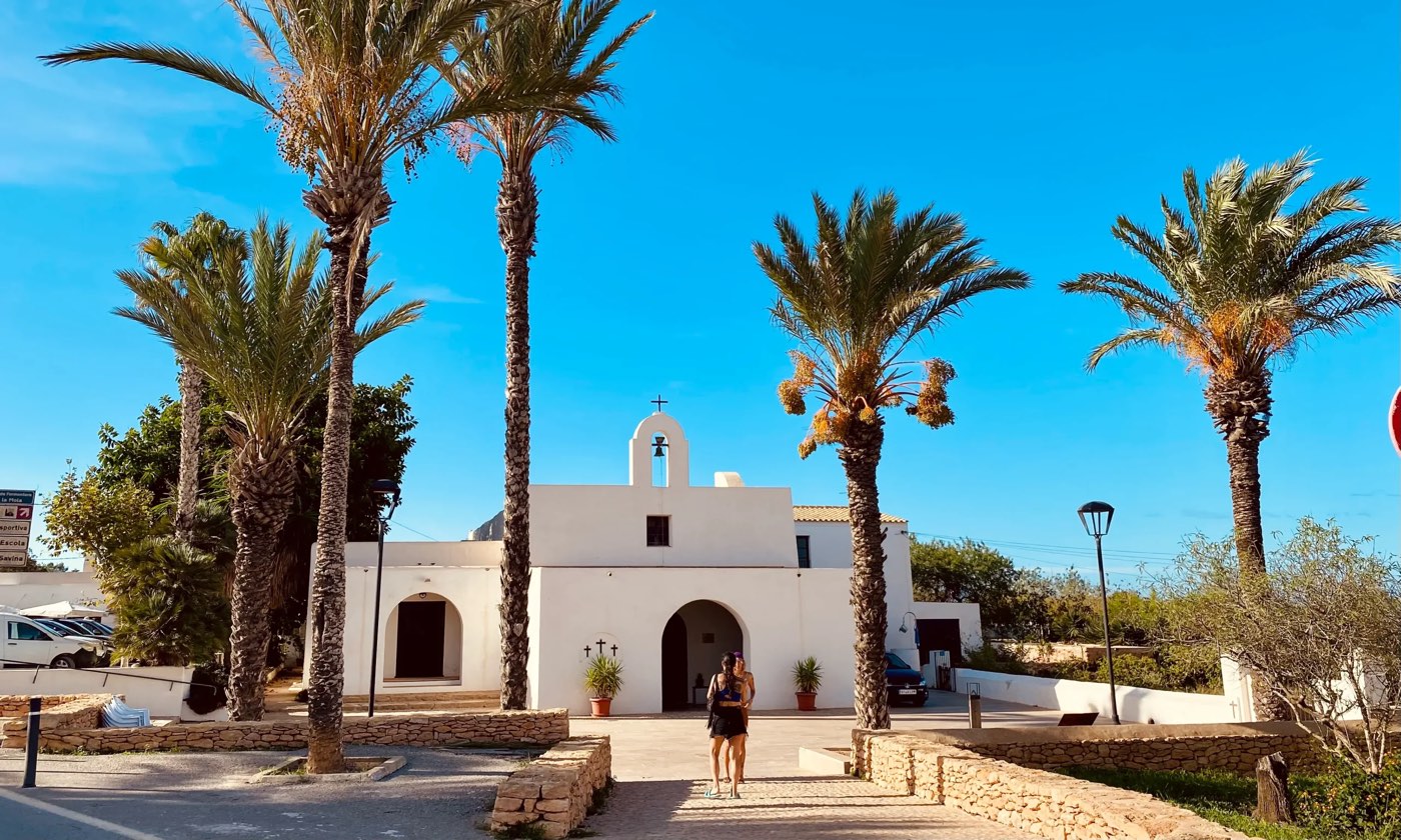 La iglesia de Nuestra Señora del Pilar de la Mola: una iglesia baja y encalada con espadaña, vista entre palmeras altas bajo un cielo azul intenso.