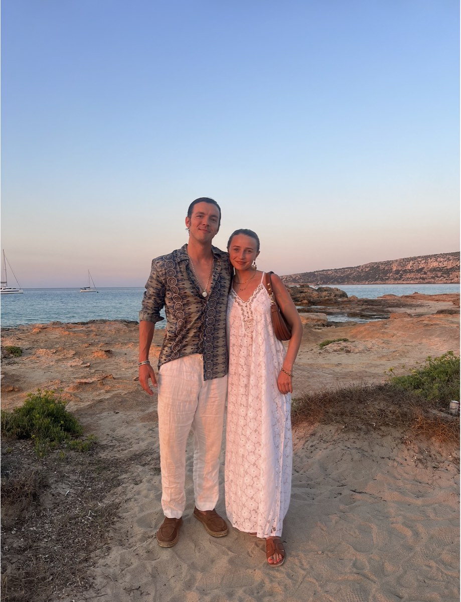 Bosco y Leah de pie sobre la arena al atardecer: &eacute;l con camisa de lino estampada abierta y pantal&oacute;n de lino blanco, ella con un vestido largo de algod&oacute;n blanco y sandalias planas.